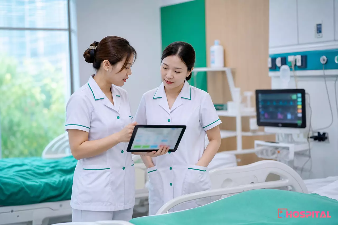 Hai điều dưỡng giao ca ở đầu giường bệnh với bản tóm tắt trên máy tính bảng