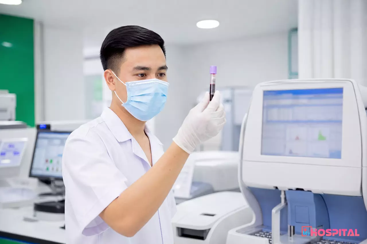 Kỹ thuật viên xét nghiệm kiểm tra ống máu có nhãn mã vạch bên cạnh máy phân tích trong phòng xét nghiệm bệnh viện
