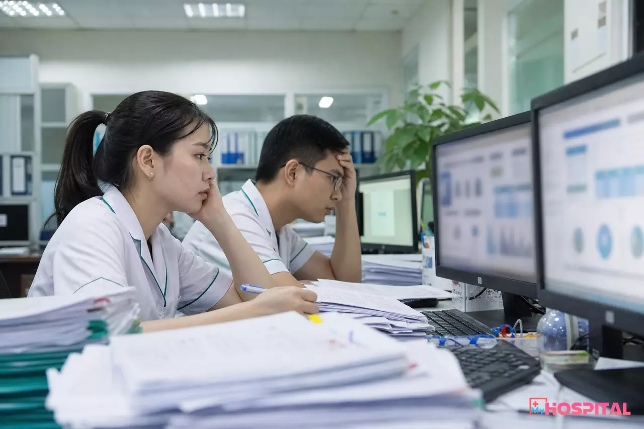 Cán bộ thống kê bệnh viện làm báo cáo cuối tháng giữa chồng giấy và nhiều bảng tính