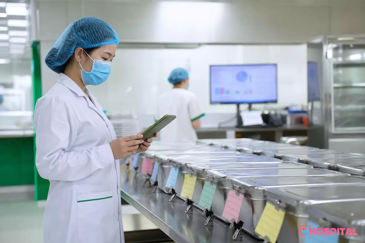 Cán bộ dinh dưỡng đối chiếu danh sách suất ăn trên máy tính bảng với các khay đã chuẩn bị