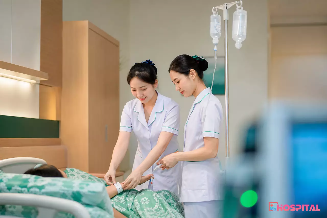 Hai điều dưỡng kiểm tra vòng đeo tay và dịch truyền của người bệnh khi bàn giao tại giường