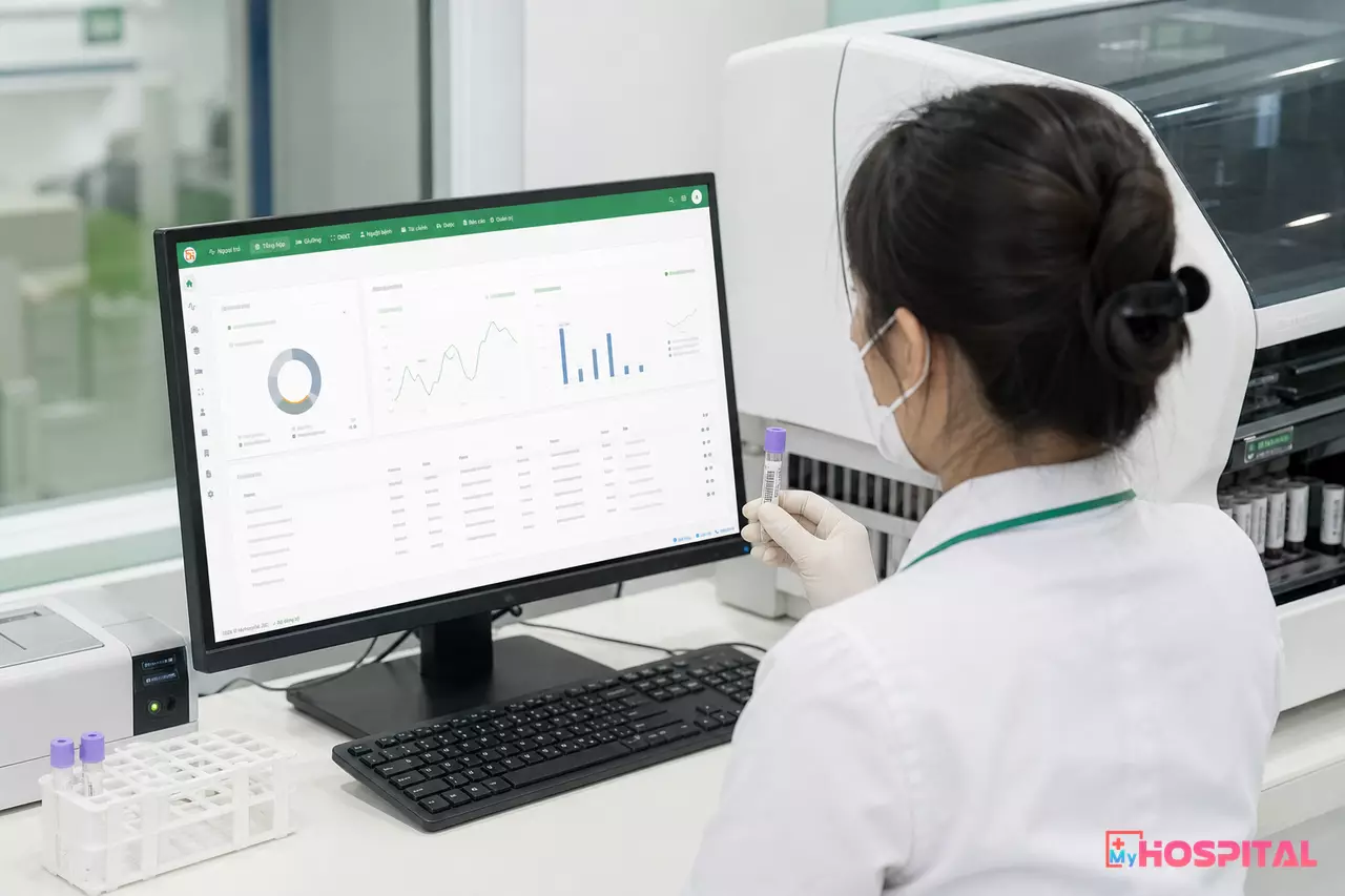 Kỹ thuật viên ngồi trước máy tính cạnh máy miễn dịch, đối chiếu kết quả với ống mẫu có mã vạch