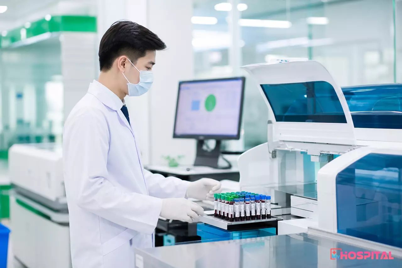 Kỹ thuật viên xét nghiệm đứng cạnh máy phân tích, ống nghiệm có nhãn mã vạch trong phòng xét nghiệm bệnh viện